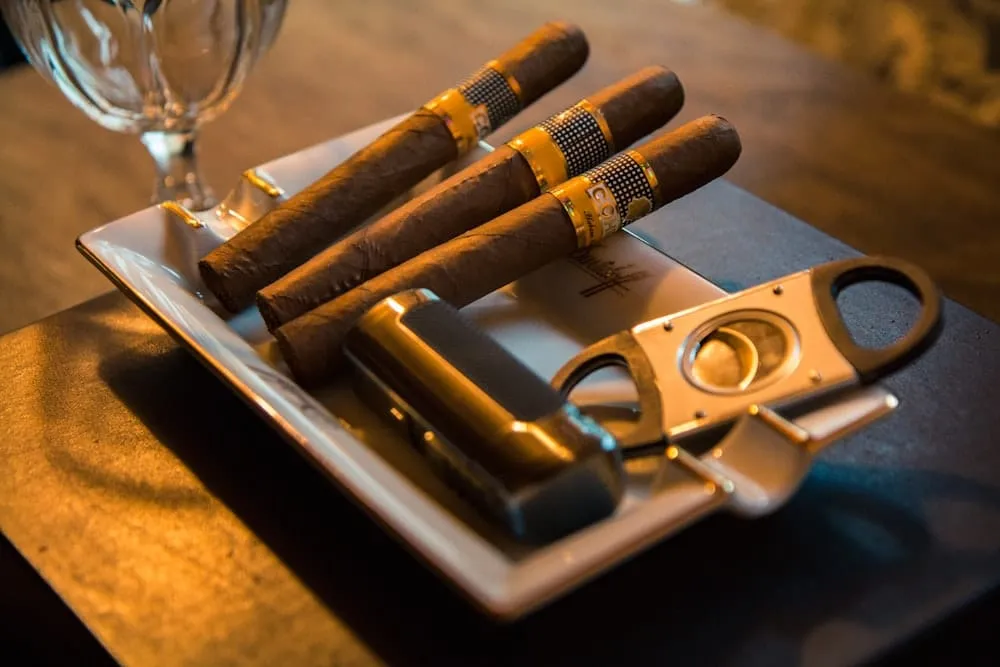 Ba điếu Cohiba vòng nhẫn ô caro trên gạt tàn trắng, cạnh bật lửa kim loại và dao cắt